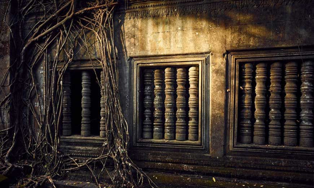 Ancient stone temple windows at Angkor overgrown with giant tree roots.